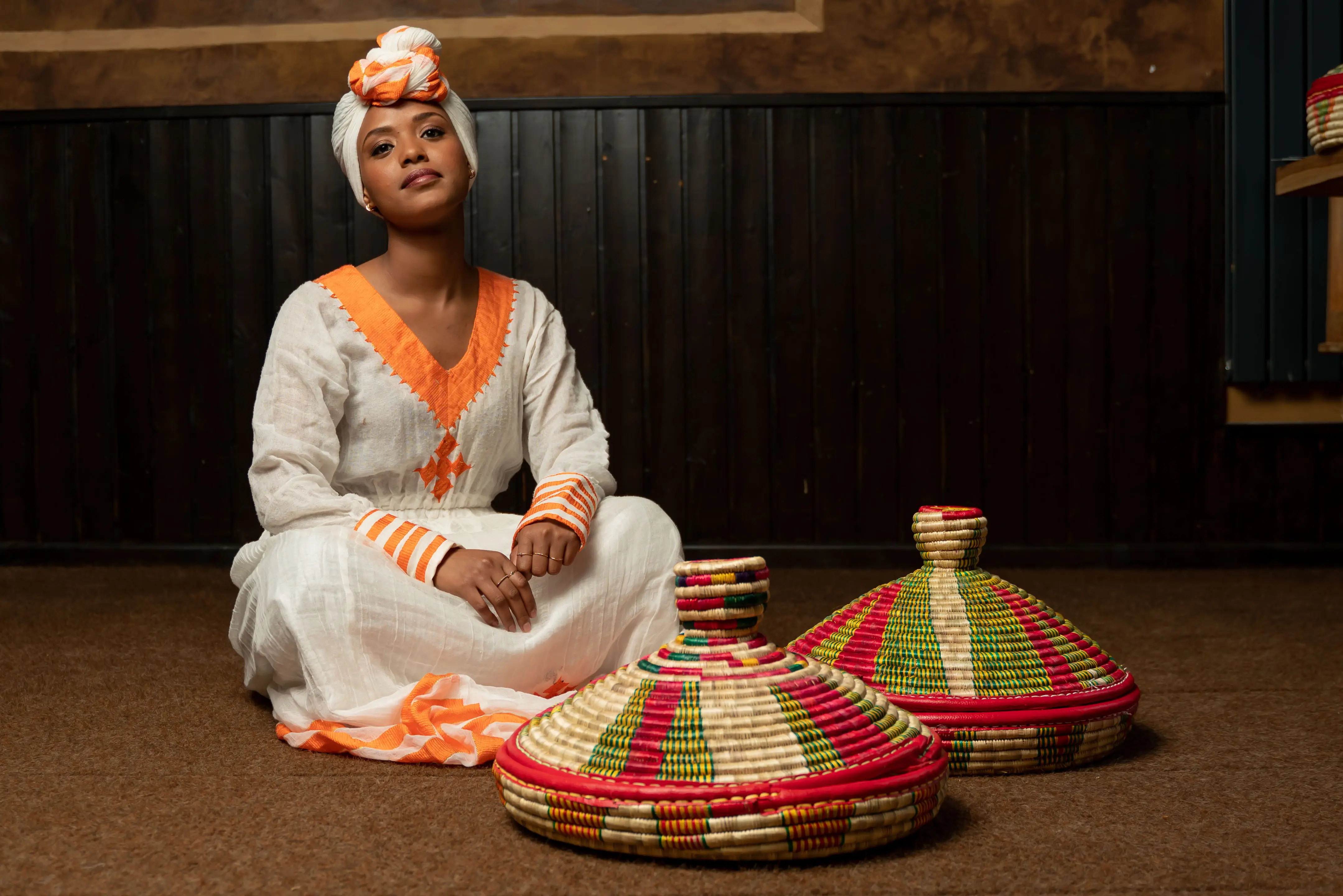 Eine Frau im traditionellen Habesha-Kemis sitzt im Lalibela neben zwei bunt geflochtenen Mesob-Körben